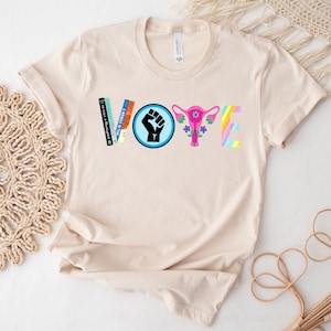 May include: A beige t-shirt with the word "VOTE" spelled out in colorful letters. The "V" is made of books, the "O" is a blue circle with a raised fist, the "T" is a pink uterus with flowers, and the "E" is a rainbow.