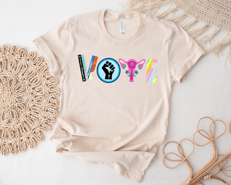 May include: A beige t-shirt with the word "VOTE" spelled out in colorful letters. The "V" is made of books, the "O" is a blue circle with a raised fist, the "T" is a pink uterus with flowers, and the "E" is a rainbow.