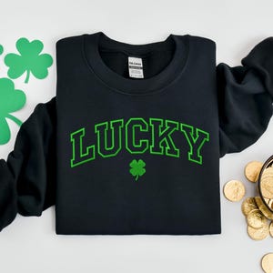 K&ouml;nnte beinhalten: Schwarzer Pullover mit gr&uuml;nem Text, der in einer College-Schriftart "LUCKY" lautet. Unter dem Text befindet sich ein gr&uuml;nes Kleeblatt.