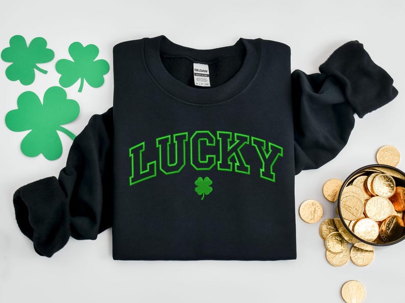 K&ouml;nnte beinhalten: Schwarzer Pullover mit gr&uuml;nem Text, der in einer College-Schriftart "LUCKY" lautet. Unter dem Text befindet sich ein gr&uuml;nes Kleeblatt.