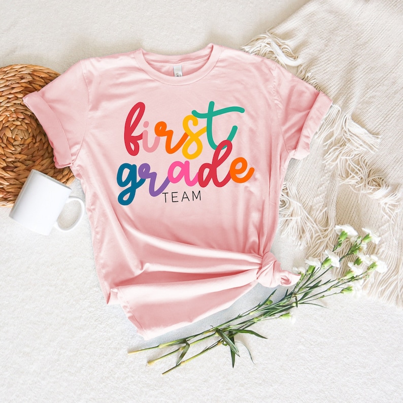K&ouml;nnte beinhalten: Rosa T-Shirt mit dem Text "first grade team" in Regenbogenfarben.