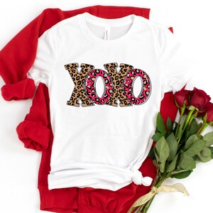 May include: White t-shirt with a pink and brown leopard print "XOXO" design. The shirt is folded and displayed with a red sweater and a bouquet of red roses.
