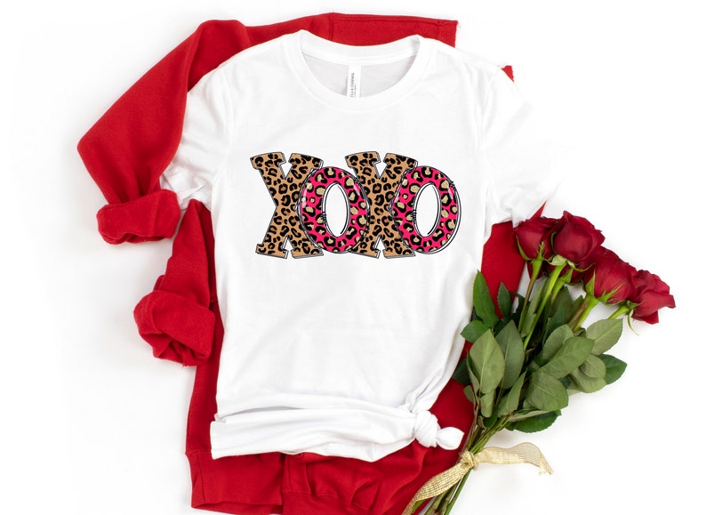 May include: White t-shirt with a pink and brown leopard print "XOXO" design. The shirt is folded and displayed with a red sweater and a bouquet of red roses.