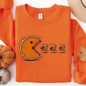 May include: An orange crewneck sweatshirt featuring a graphic print of a pumpkin pie slice shaped like a video game character chasing three brown turkeys. The design includes motion lines behind the turkeys and a white brand tag at the neck.