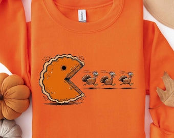 Puede incluir: Una sudadera naranja de cuello redondo con un estampado gráfico de un trozo de tarta de calabaza con forma de personaje de videojuego persiguiendo a tres pavos marrones. El diseño incluye líneas de movimiento y una etiqueta blanca.