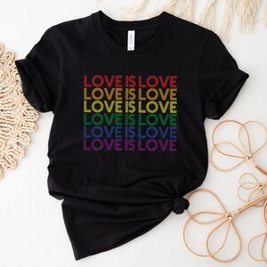 Może przedstawiać: Czarny T-shirt z napisem "LOVE IS LOVE" powt&oacute;rzonym sześć razy w kolorach tęczy.