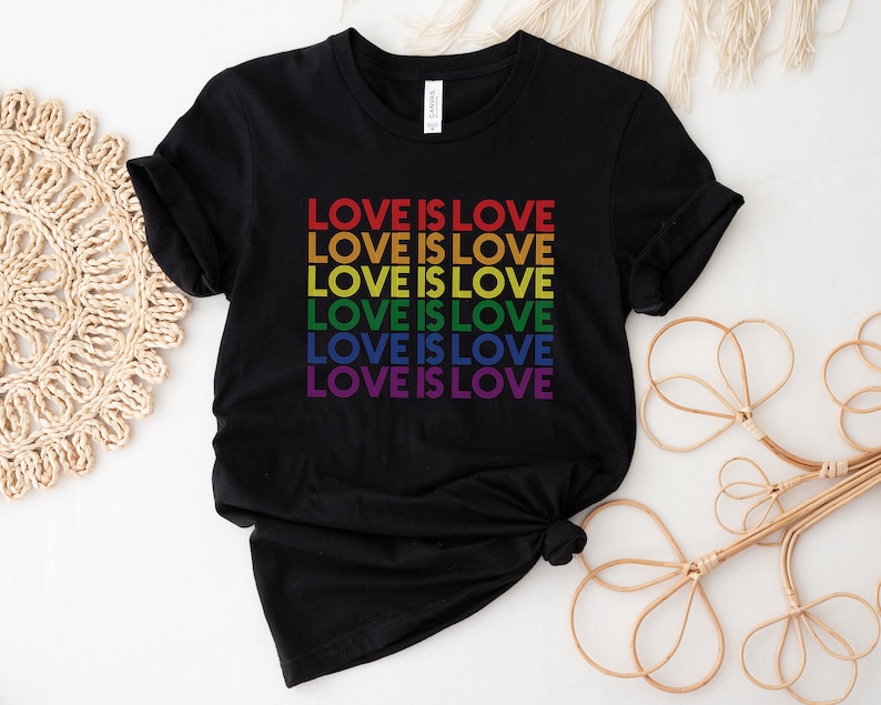 Może przedstawiać: Czarny T-shirt z napisem "LOVE IS LOVE" powt&oacute;rzonym sześć razy w kolorach tęczy.