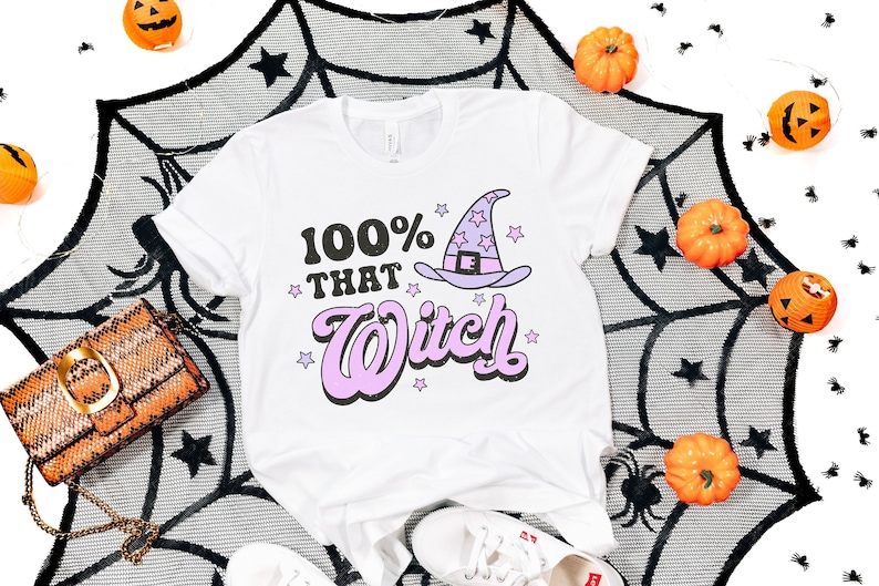 K&ouml;nnte beinhalten: Wei&szlig;es T-Shirt mit einem lila Hexenhut und dem Text "100% That Witch" in schwarzen und pinken Buchstaben. Das Shirt ist von einem schwarzen Spinnennetz und orangefarbenen K&uuml;rbissen umgeben.