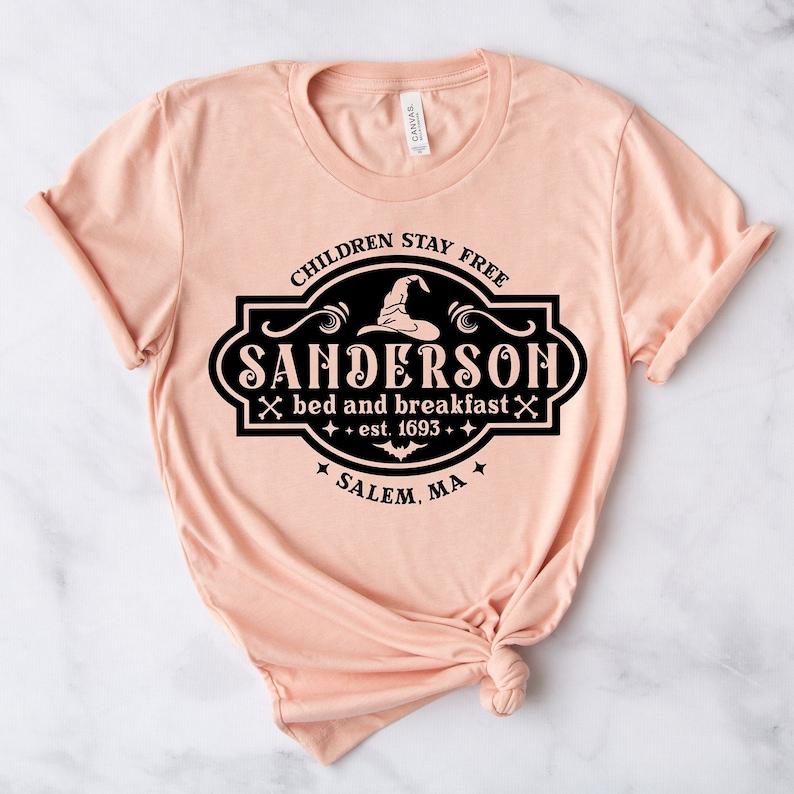 May include: A light peach-colored t-shirt with a black graphic design. The design features the text "CHILDREN STAY FREE" above "SANDERSON bed and breakfast est. 1693" with a witch hat and bat illustration. The text "SALEM, MA" is at the bottom.