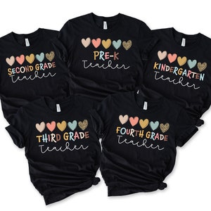 K&ouml;nnte beinhalten: F&uuml;nf schwarze T-Shirts mit bunten Herzen und dem Text "SECOND GRADE Teacher", "PRE-K Teacher", "KINDERGARTEN Teacher", "THIRD GRADE Teacher" und "FOURTH GRADE Teacher" bedruckt.