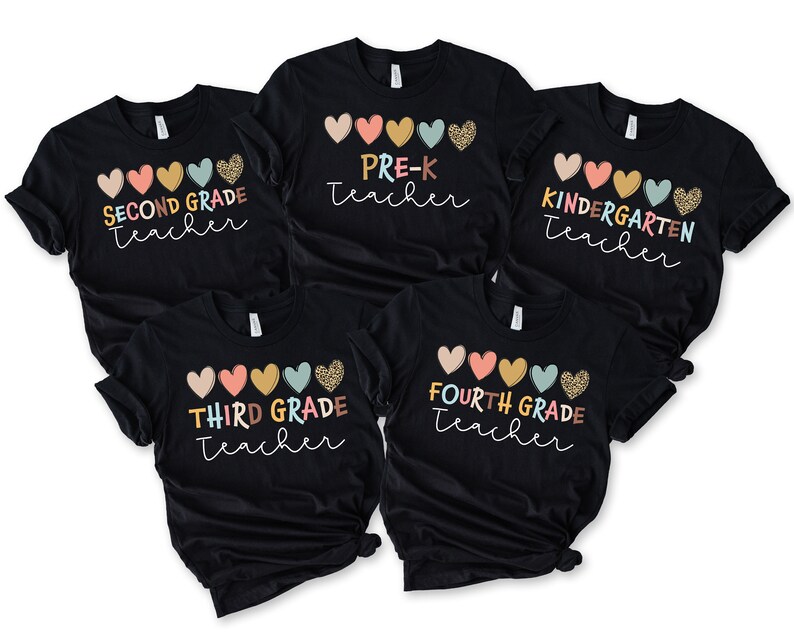 K&ouml;nnte beinhalten: F&uuml;nf schwarze T-Shirts mit bunten Herzen und dem Text "SECOND GRADE Teacher", "PRE-K Teacher", "KINDERGARTEN Teacher", "THIRD GRADE Teacher" und "FOURTH GRADE Teacher" bedruckt.