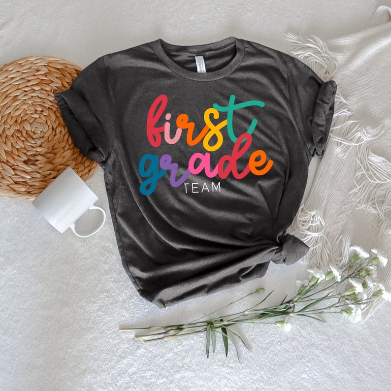 K&ouml;nnte beinhalten: Ein dunkelgraues T-Shirt mit den Worten "first grade team" in Regenbogenfarben.