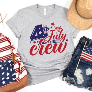 Op de afbeelding: Een grijs T-shirt met de tekst "4th of July Crew" in rood, wit en blauw. De tekst is in een feestelijk lettertype en bevat sterren en strepen.