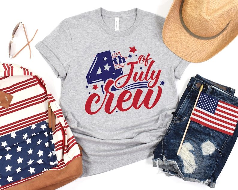 Op de afbeelding: Een grijs T-shirt met de tekst "4th of July Crew" in rood, wit en blauw. De tekst is in een feestelijk lettertype en bevat sterren en strepen.