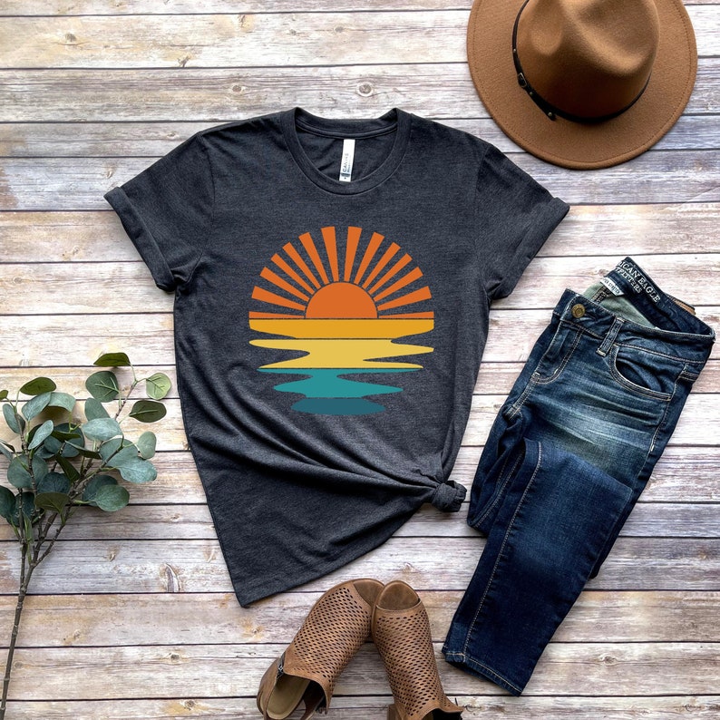 May include: A dark gray t-shirt with a retro sunset graphic featuring a yellow sun with orange rays over a blue and green ocean. The shirt is laid flat on a wooden surface with a pair of brown sandals, blue jeans, and a brown fedora hat.