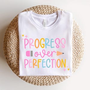 May include: White t-shirt with the phrase "PROGRESS OVER PERFECTION" in colorful, playful lettering. The design includes a pencil graphic and star accents. The shirt is displayed on a woven, circular surface.