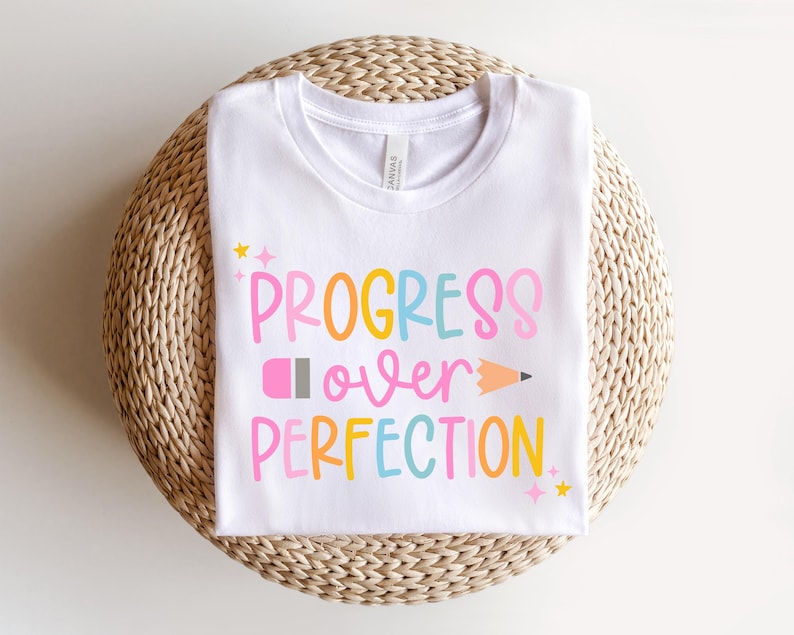 May include: White t-shirt with the phrase "PROGRESS OVER PERFECTION" in colorful, playful lettering. The design includes a pencil graphic and star accents. The shirt is displayed on a woven, circular surface.