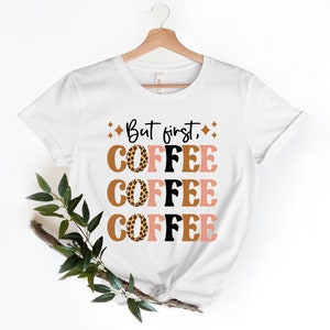 May include: White t-shirt with the text "But first, coffee" printed in black. The word "coffee" is repeated three times, each time with a different colored background and a leopard print design.