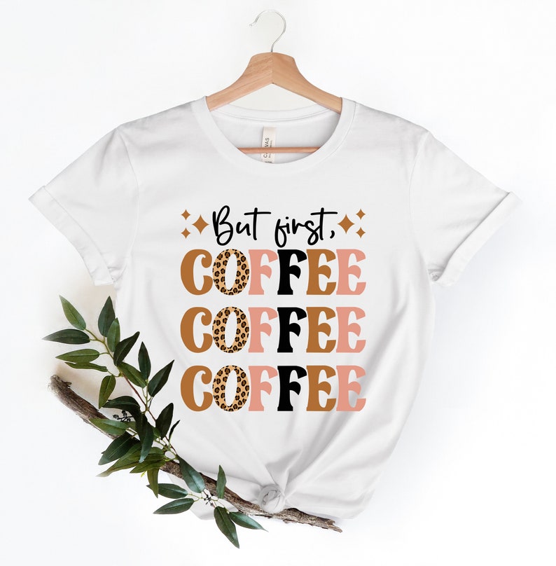 May include: White t-shirt with the text "But first, coffee" printed in black. The word "coffee" is repeated three times, each time with a different colored background and a leopard print design.