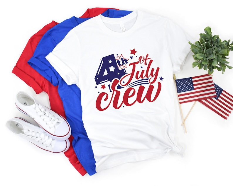 Op de afbeelding: Wit T-shirt met een rood, wit en blauwe afbeelding met de tekst "4th of July Crew". Het shirt is gevouwen en wordt getoond met rode en blauwe T-shirts eronder.