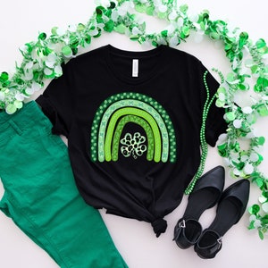 May include: A black t-shirt with a green shamrock rainbow design. The shirt is laid out on a white surface with green shamrock garland and green pants.