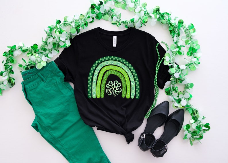 May include: A black t-shirt with a green shamrock rainbow design. The shirt is laid out on a white surface with green shamrock garland and green pants.