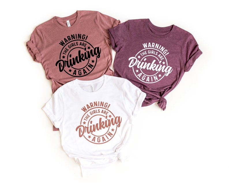 May include: Three t-shirts in shades of mauve, burgundy, and white, each featuring the text "WARNING! THE GIRLS ARE Drinking AGAIN" in a circular design. The text is in black, white, and a copper color.