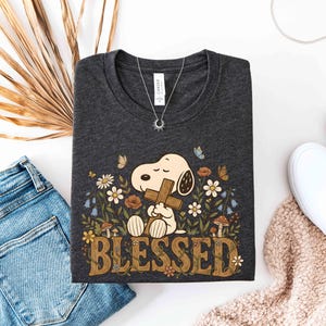 Puede incluir: Camiseta gris oscuro con Snoopy sosteniendo una cruz, rodeado de flores y mariposas, con la palabra "BLESSED". Tambi&eacute;n se ven un collar plateado y zapatos blancos. La camiseta est&aacute; doblada.