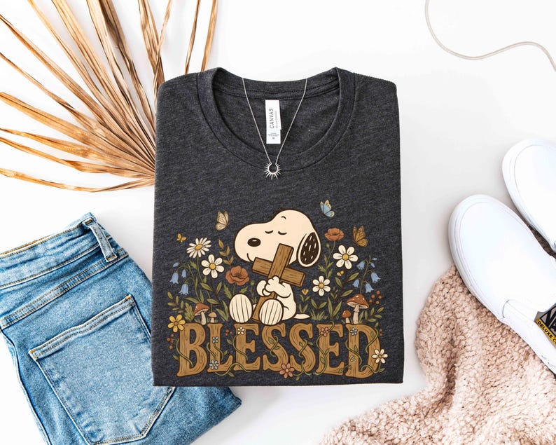 Puede incluir: Camiseta gris oscuro con Snoopy sosteniendo una cruz, rodeado de flores y mariposas, con la palabra "BLESSED". Tambi&eacute;n se ven un collar plateado y zapatos blancos. La camiseta est&aacute; doblada.