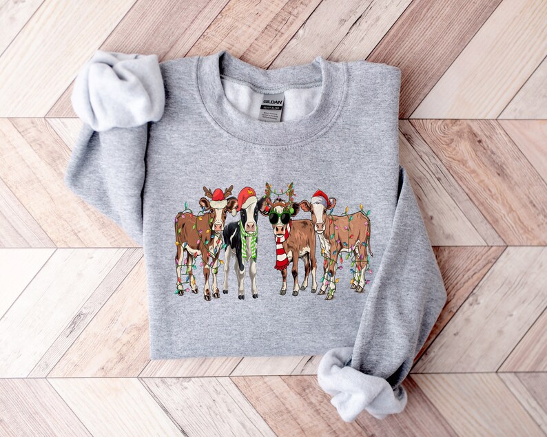 May include: A gray sweatshirt with a Christmas-themed design featuring four cows wearing Santa hats and reindeer antlers. The cows are decorated with string lights.