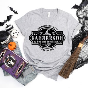 May include: A heather gray t-shirt with a black and white Sanderson Bed and Breakfast design. The design includes a witch hat, crossed broomsticks, and the text "Children Stay Free" and "Salem, MA". Halloween-themed apparel.