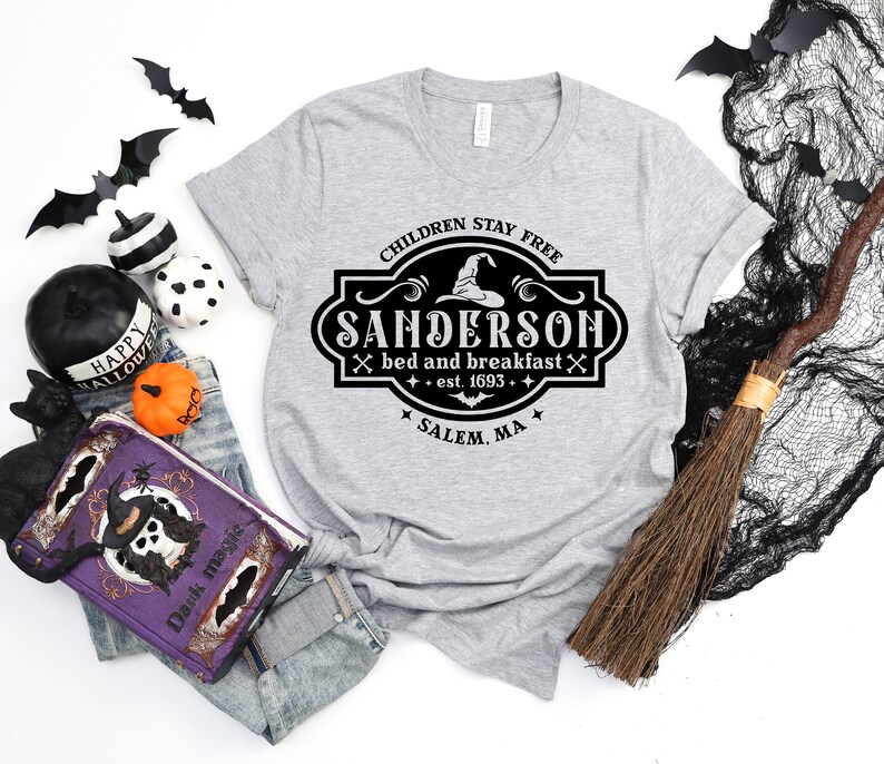 May include: A heather gray t-shirt with a black and white Sanderson Bed and Breakfast design. The design includes a witch hat, crossed broomsticks, and the text "Children Stay Free" and "Salem, MA". Halloween-themed apparel.