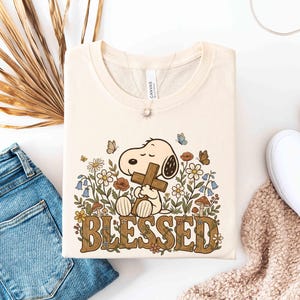 Puede incluir: Camiseta color crema con un dibujo de Snoopy sosteniendo una cruz, rodeado de flores y mariposas. La palabra "BLESSED" est&aacute; impresa en marr&oacute;n con un dise&ntilde;o floral. La camiseta est&aacute; doblada sobre una superficie blanca, junto a unos vaqueros azules y zapatos blancos.