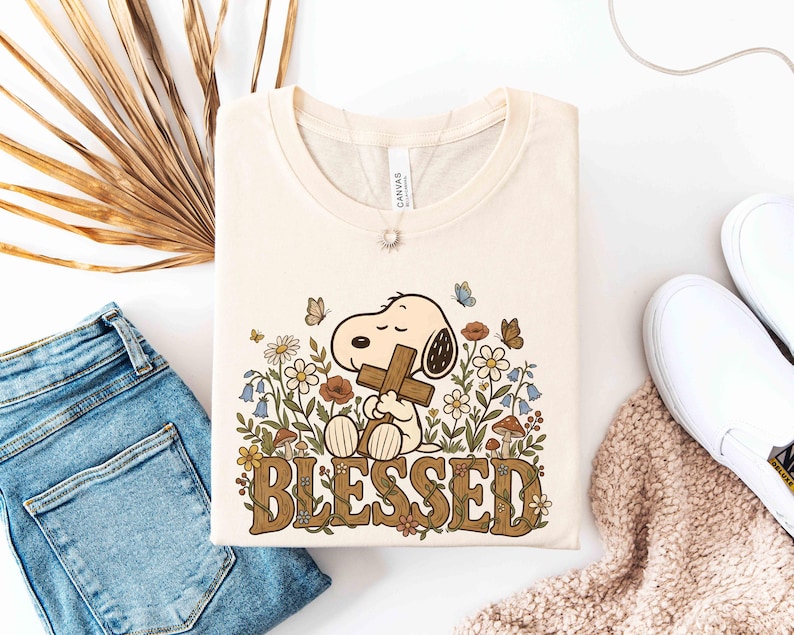 Puede incluir: Camiseta color crema con un dibujo de Snoopy sosteniendo una cruz, rodeado de flores y mariposas. La palabra "BLESSED" est&aacute; impresa en marr&oacute;n con un dise&ntilde;o floral. La camiseta est&aacute; doblada sobre una superficie blanca, junto a unos vaqueros azules y zapatos blancos.