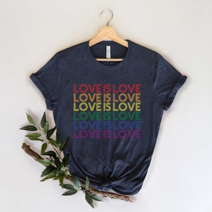 Może przedstawiać: Koszulka w kolorze ciemnoniebieskim z napisem "LOVE IS LOVE" powt&oacute;rzonym sześć razy w kolorach tęczy.
