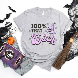 K&ouml;nnte beinhalten: Ein graues T-Shirt mit dem Text "100% That Witch" in rosa und schwarzen Buchstaben. Ein Cartoon-Hexenhut mit Sternen befindet sich &uuml;ber dem Text.