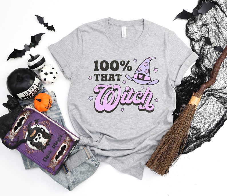 K&ouml;nnte beinhalten: Ein graues T-Shirt mit dem Text "100% That Witch" in rosa und schwarzen Buchstaben. Ein Cartoon-Hexenhut mit Sternen befindet sich &uuml;ber dem Text.