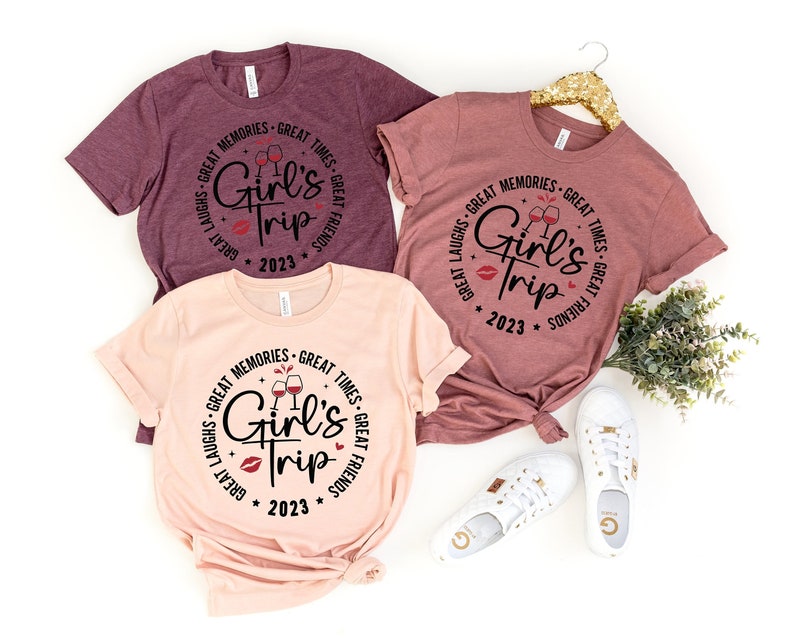 K&ouml;nnte beinhalten: Drei T-Shirts mit einem kreisf&ouml;rmigen Design mit dem Text "Girl's Trip" mit Weingl&auml;sern und dem Jahr 2023. Die Shirts sind in Rosat&ouml;nen und Rotbraun.