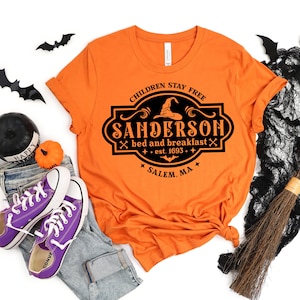 May include: Orange t-shirt with a black graphic that reads "CHILDREN STAY FREE SANDERSON bed and breakfast est. 1693 SALEM, MA." The shirt is styled with jeans, purple sneakers, and Halloween decorations.