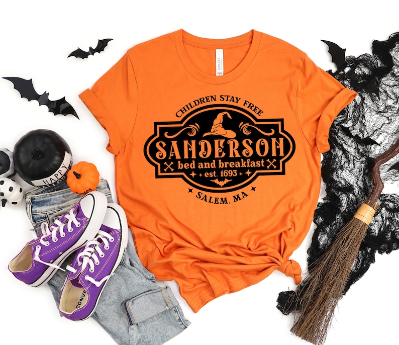 May include: Orange t-shirt with a black graphic that reads "CHILDREN STAY FREE SANDERSON bed and breakfast est. 1693 SALEM, MA." The shirt is styled with jeans, purple sneakers, and Halloween decorations.