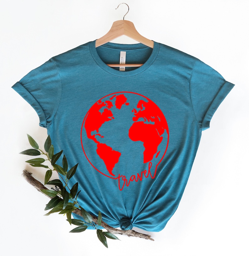 Peut inclure: T-shirt bleu sarcelle avec un globe terrestre rouge et le mot "travel" &eacute;crit en cursive rouge en dessous.