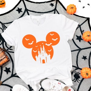 May include: White t-shirt with an orange Mickey Mouse-shaped design featuring bats and a castle silhouette. The shirt is decorated with Halloween-themed elements, including pumpkins and a spiderweb.