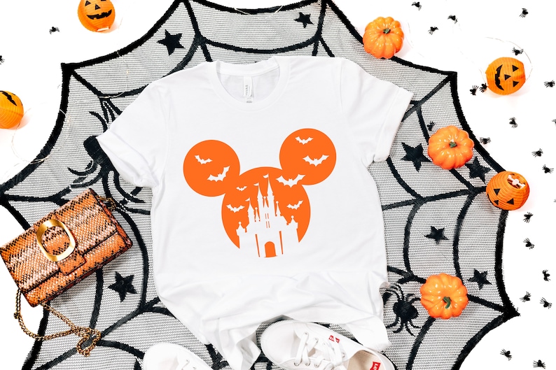 May include: White t-shirt with an orange Mickey Mouse-shaped design featuring bats and a castle silhouette. The shirt is decorated with Halloween-themed elements, including pumpkins and a spiderweb.