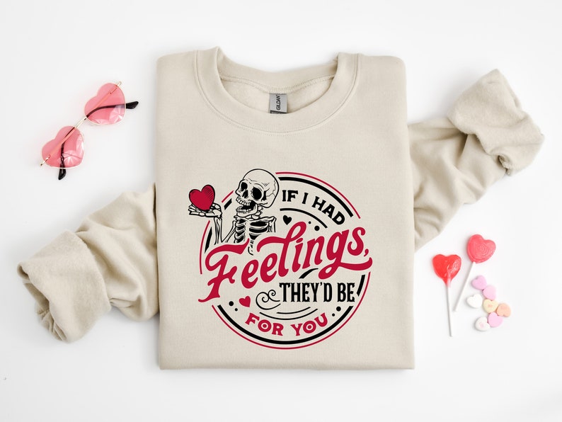 May include: A beige sweatshirt featuring a graphic print of a skeleton holding a heart. The graphic reads "If I had feelings, they'd be for you".