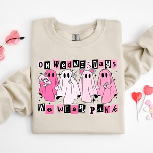May include: A beige crewneck sweatshirt featuring a graphic of four ghosts in pink and white. The text reads ON WEDNESDAYS WE WEAR PINK. One ghost holds a book labelled BOO BOOK, another holds a phone, and one wears mouse ears. The design includes black stars.