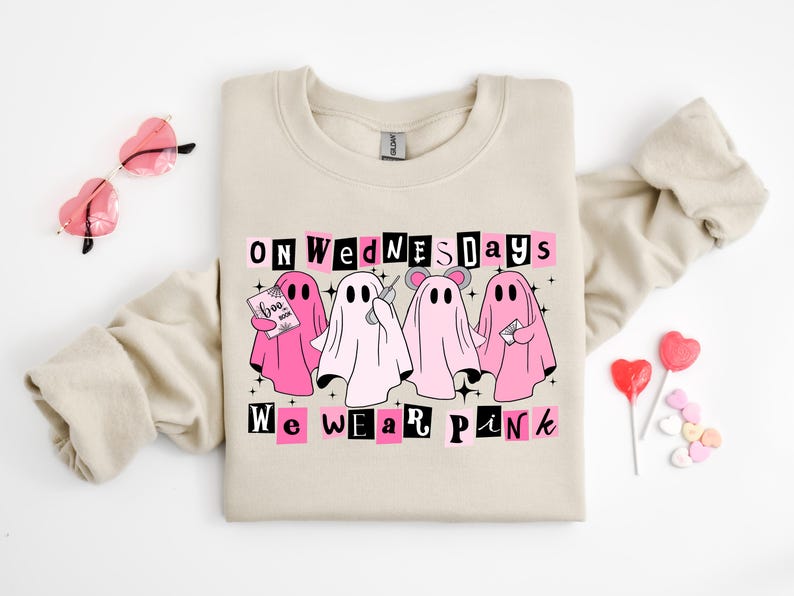 May include: A beige crewneck sweatshirt featuring a graphic of four ghosts in pink and white. The text reads ON WEDNESDAYS WE WEAR PINK. One ghost holds a book labelled BOO BOOK, another holds a phone, and one wears mouse ears. The design includes black stars.