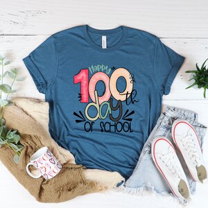 May include: A blue t-shirt with the text "Happy 100th day of school" in colorful, stylized lettering.