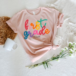 K&ouml;nnte beinhalten: Ein rosa T-Shirt mit den Worten "first grade team" in bunten, stilisierten Buchstaben.