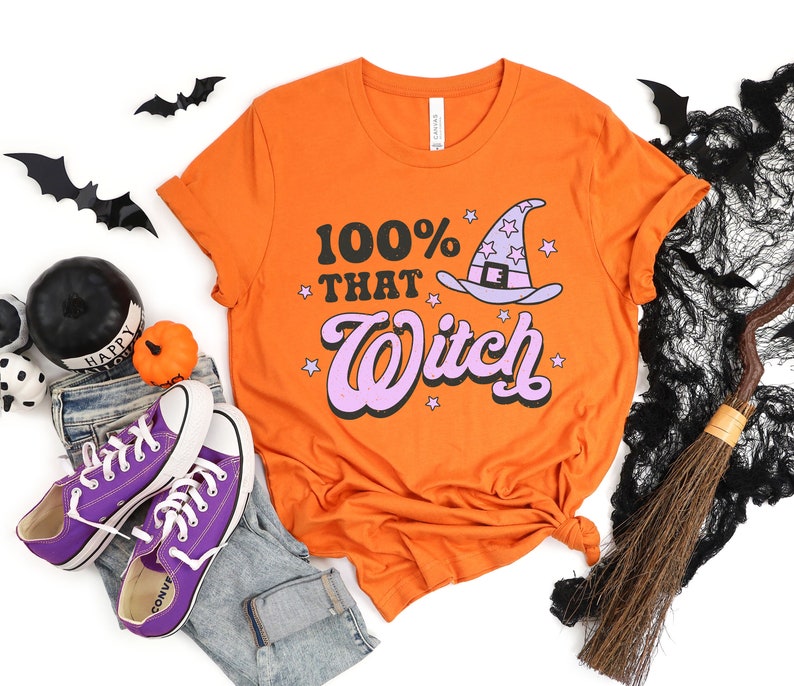 K&ouml;nnte beinhalten: Orangefarbenes T-Shirt mit einem pinkfarbenen und wei&szlig;en Hexenhut-Motiv und dem Text "100% That Witch".