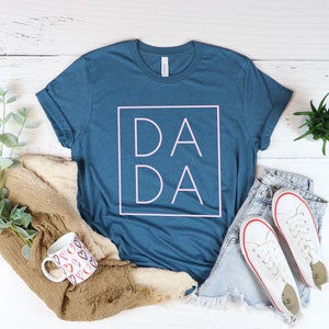 May include: A blue t-shirt with the word "DADA" in a pink square. The shirt is styled with a pair of denim shorts, white sneakers, a mug with heart designs, and a folded brown sweater. The items are arranged on a white wooden surface.